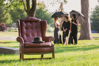 We are outdoors. In the foreground is a leather chair with a men's hat sitting on it. In the background are three women in black holding umbrellas and talking.
