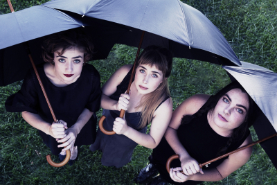 Three women dressed in funeral black standing on the grass, looking up from under black umbrellas