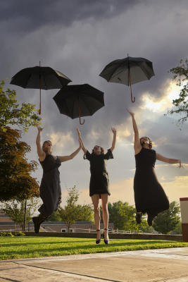Three women in funeral black clothing are tossing their umbrellas into the air and jumping to catch them