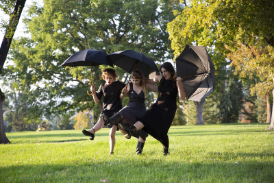 Three women are outdoors, wearing funeral black clothing, and dancing with each other while holding umbrellas