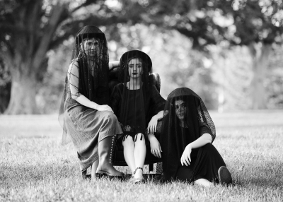 Three women in funeral clothing are sitting on a leather chair in the middle of a grassy field. They are wearing hats with black veils and staring into the camera.